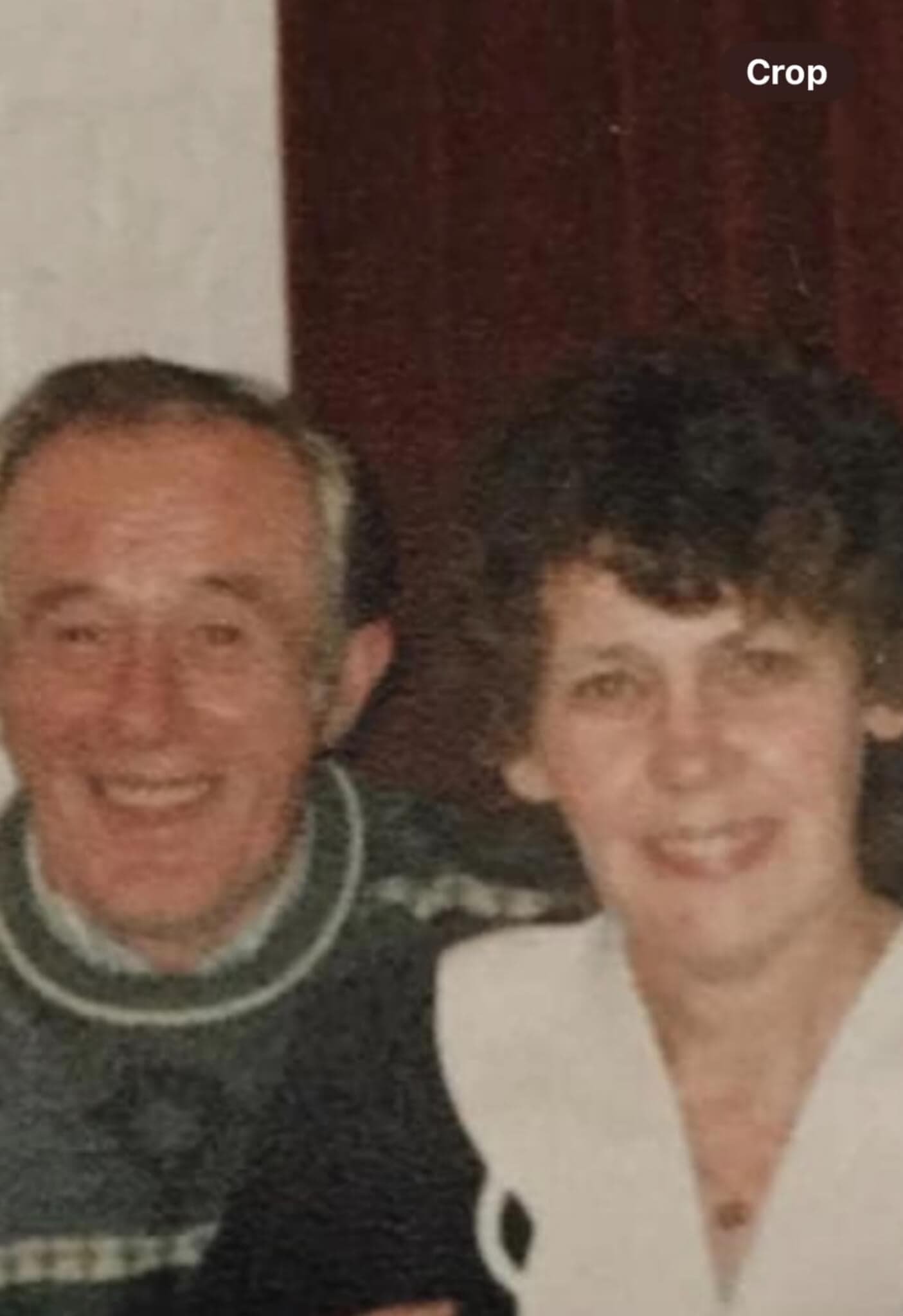 A smiling elderly man in a sweater and a woman with curly hair are posing together in front of a dark red curtain. - Home Instead