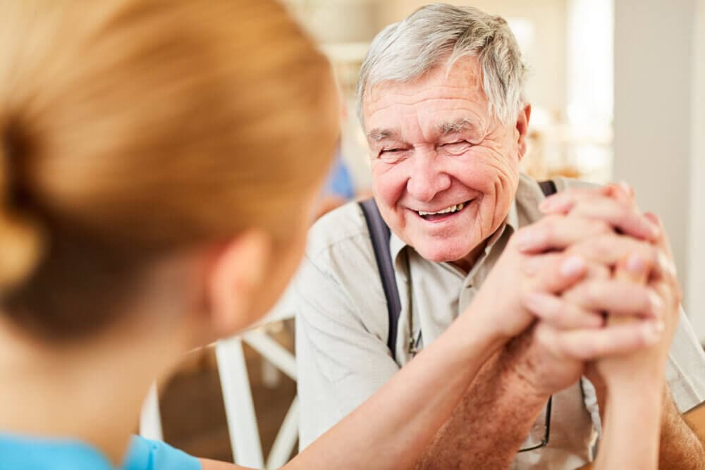 A smiling elderly man holding hands with a caregiver, creating a warm and joyful moment. - Home Instead