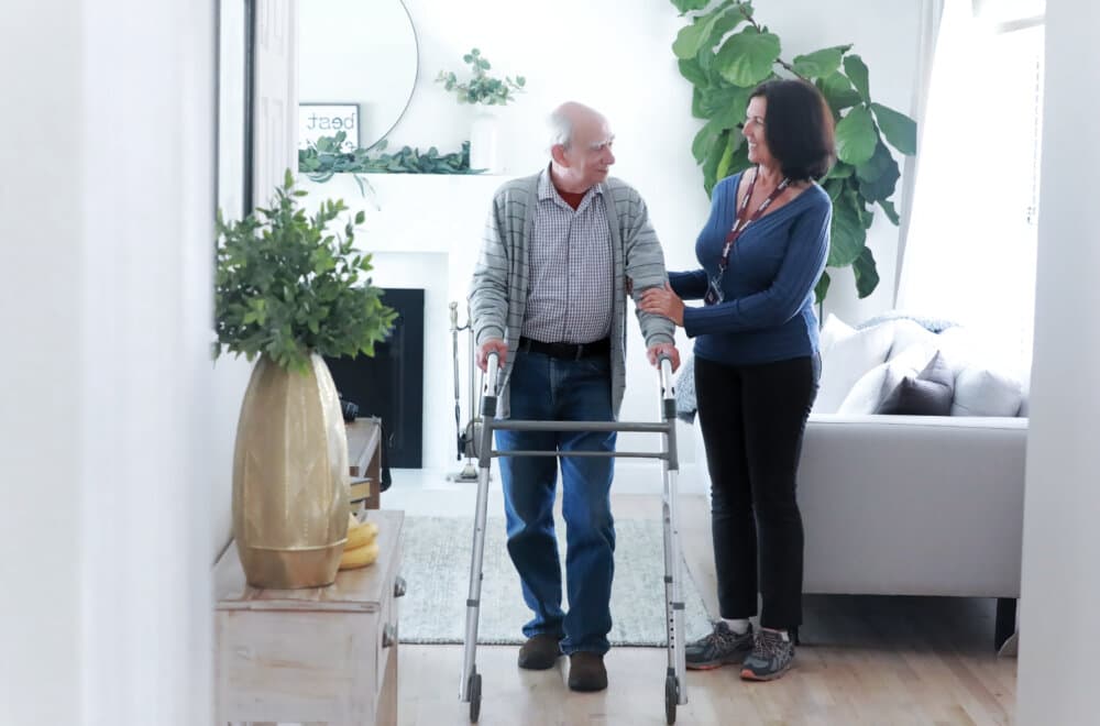 An elderly man using a walker is assisted by a woman in a living room with bright natural light and plants. - Home Instead