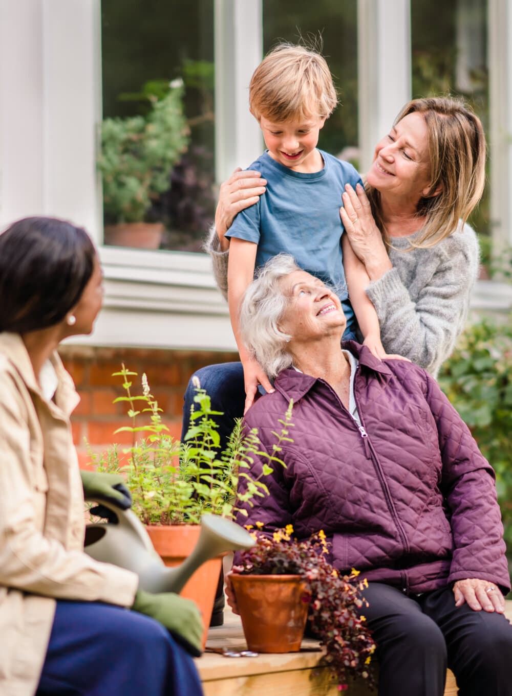 A family of three generations enjoys time together in a garden, including two women, one boy, and potted plants. - Home Instead