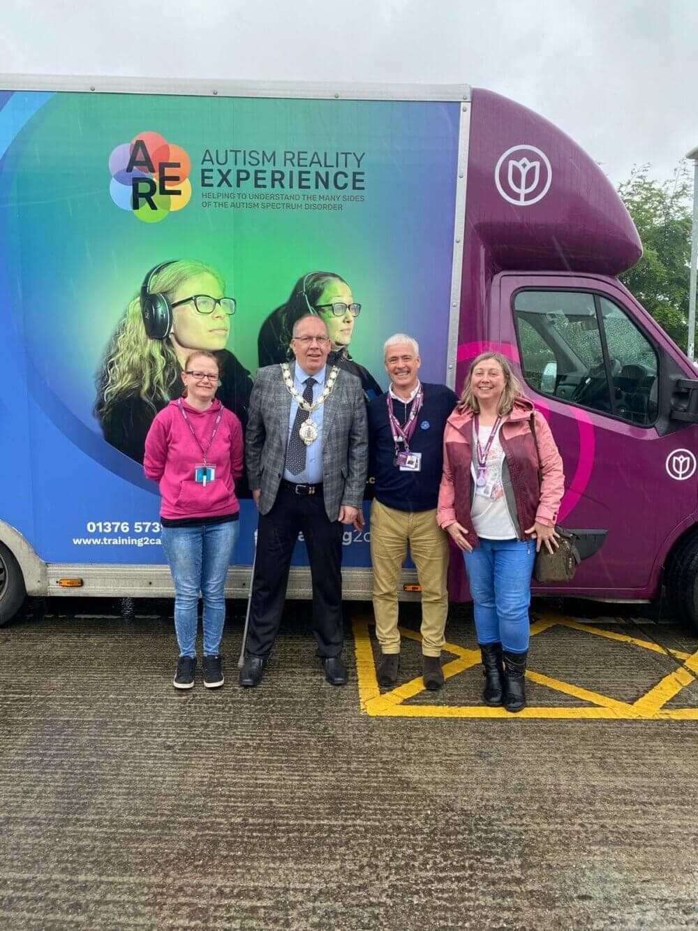 Four people stand in front of an Autism Reality Experience vehicle. The man in the middle wears a chain of office. - Home Instead