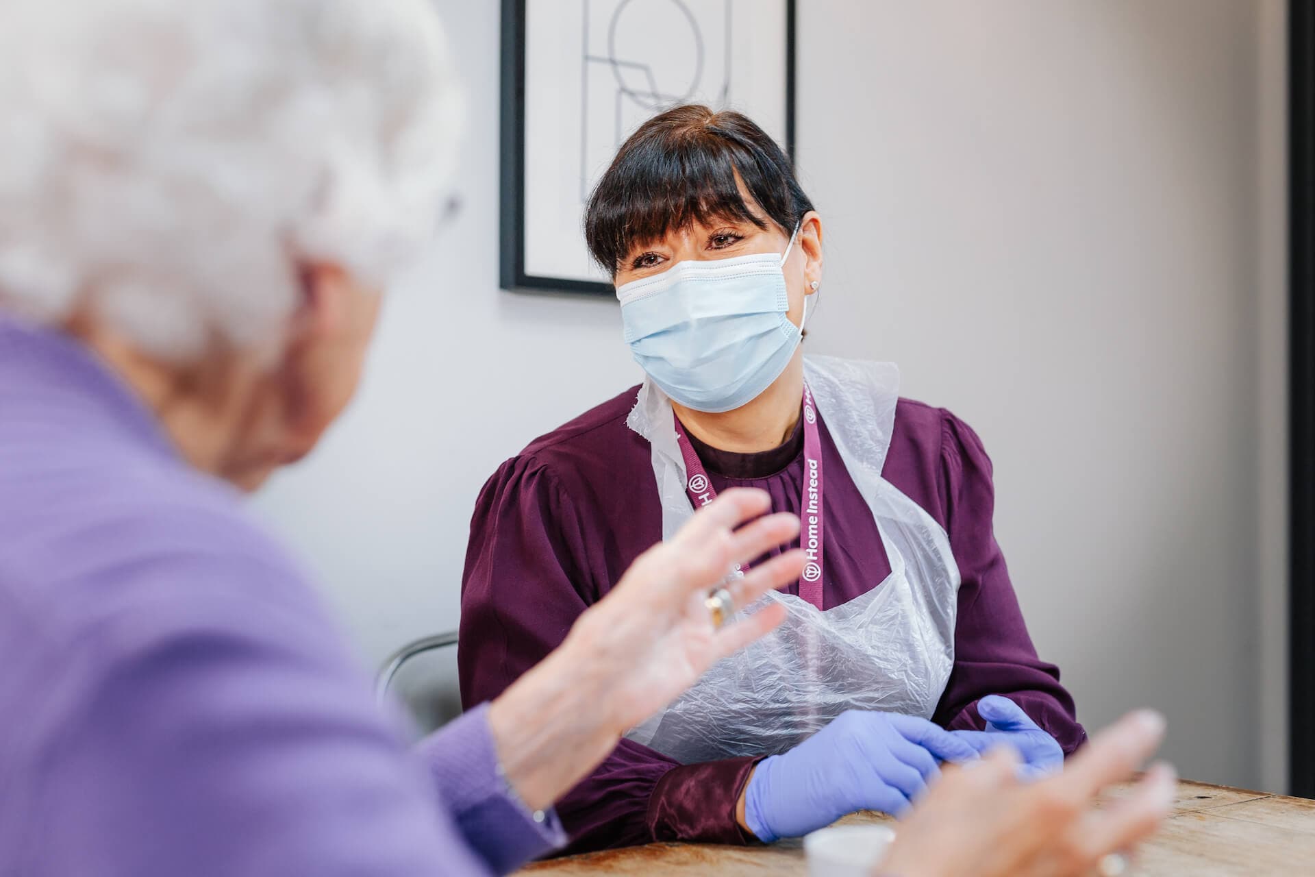 A healthcare worker wearing a mask and gloves talks to an elderly woman in a purple sweater. - Home Instead