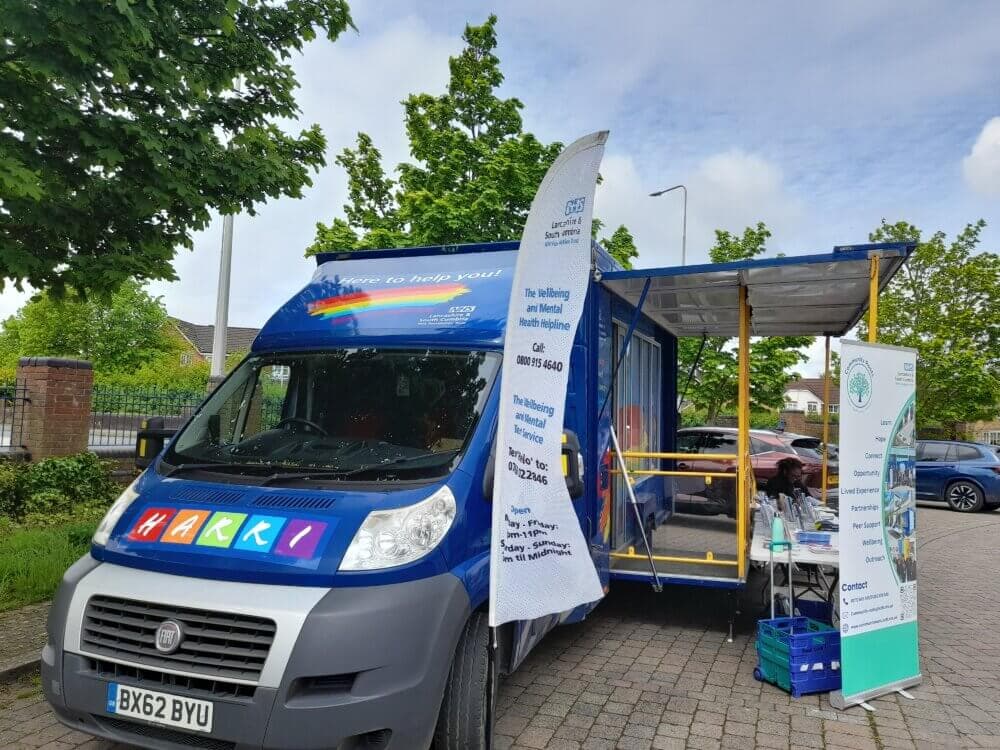 A blue mental health support van with informational banners is parked outdoors on a paved area with trees nearby. - Home Instead