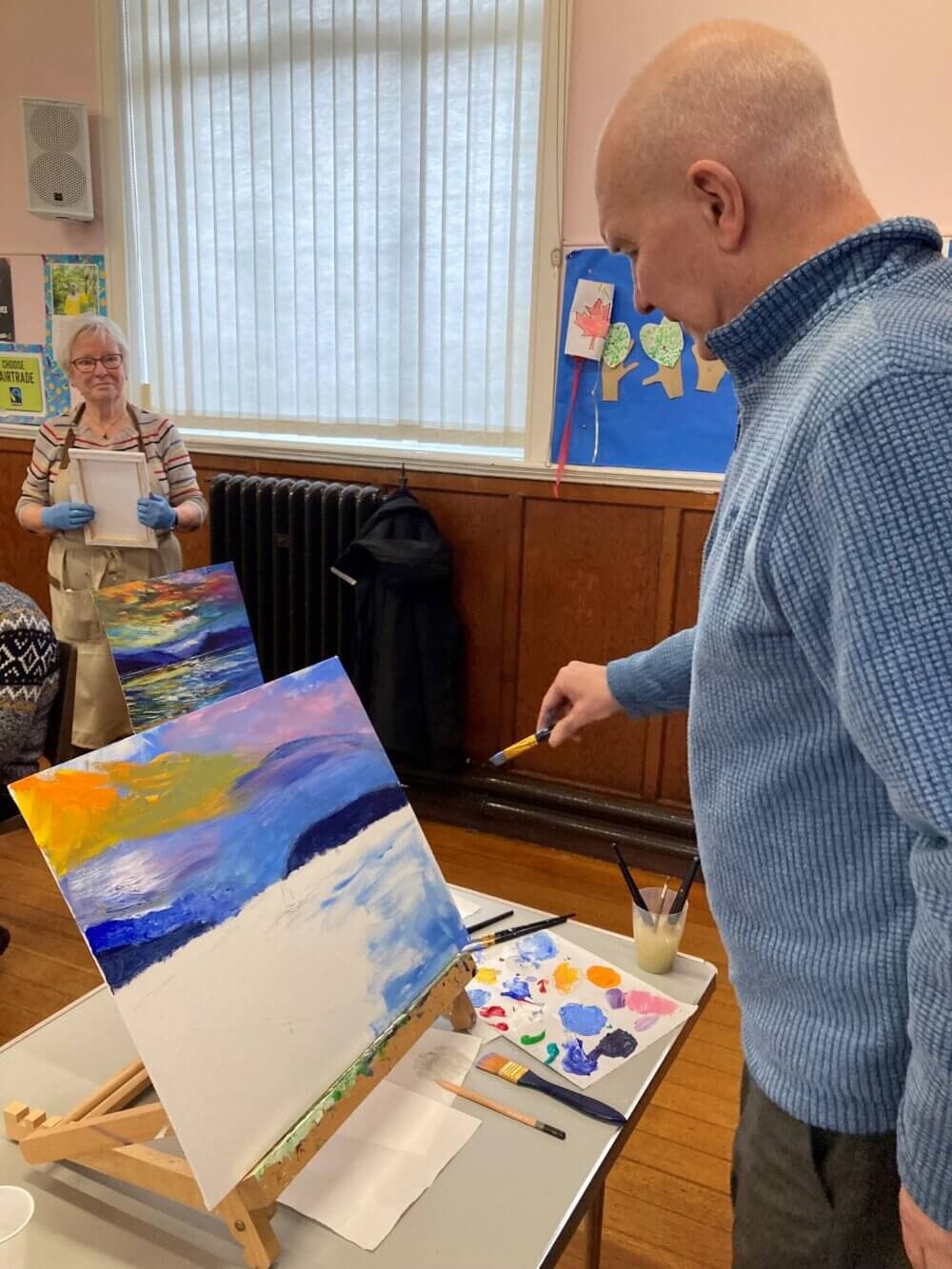 A man in a blue sweater paints a colorful landscape, while a woman in overalls smiles and observes in the background. - Home Instead