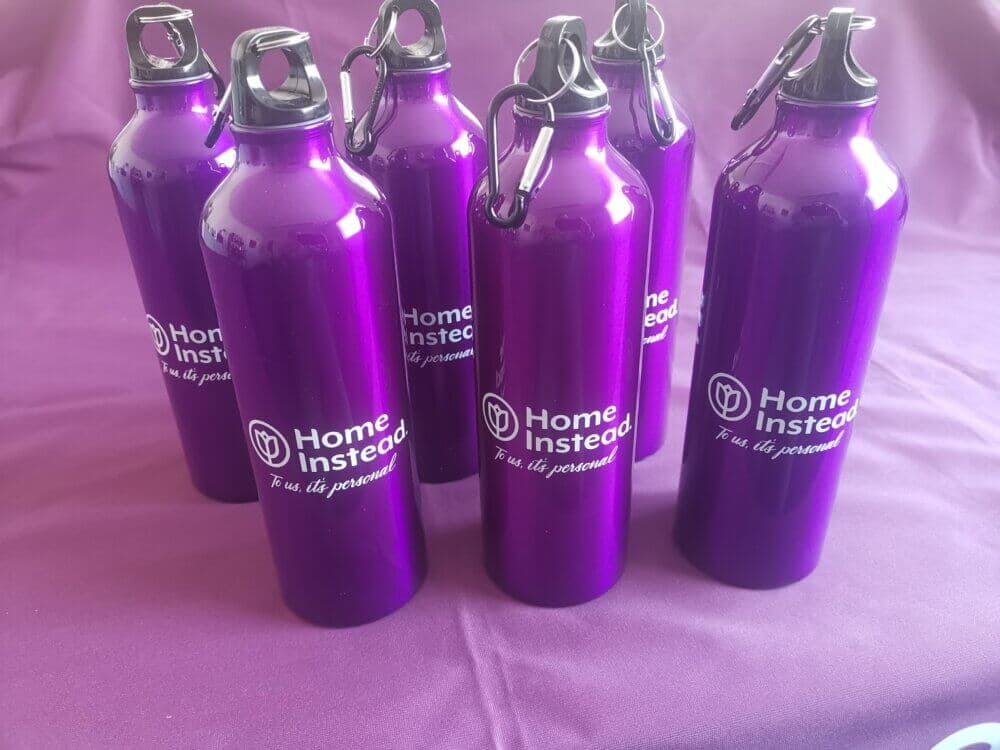 Seven purple "Home Instead" branded water bottles arranged on a purple surface. - Home Instead