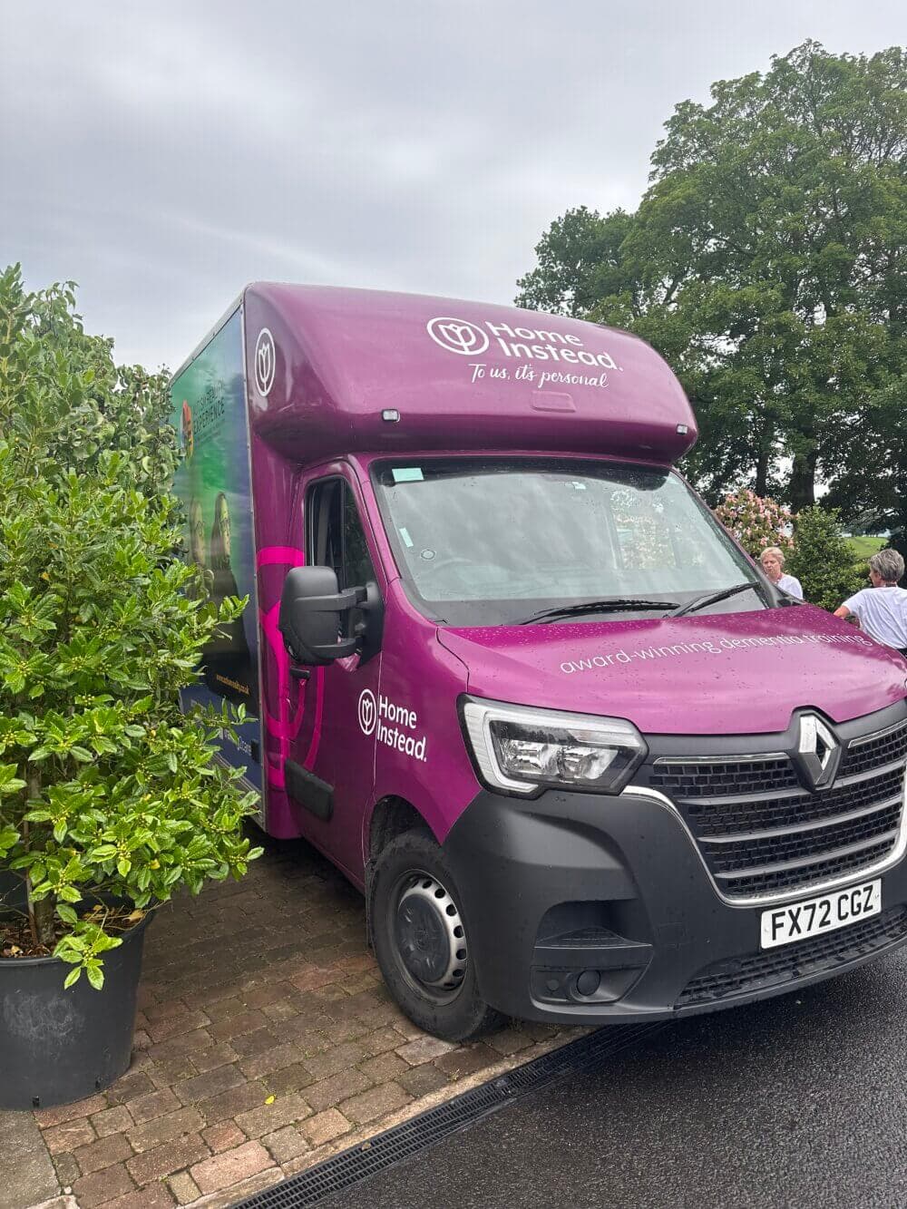 A purple Home Instead delivery van is parked on a driveway, surrounded by greenery and trees in the background. - Home Instead