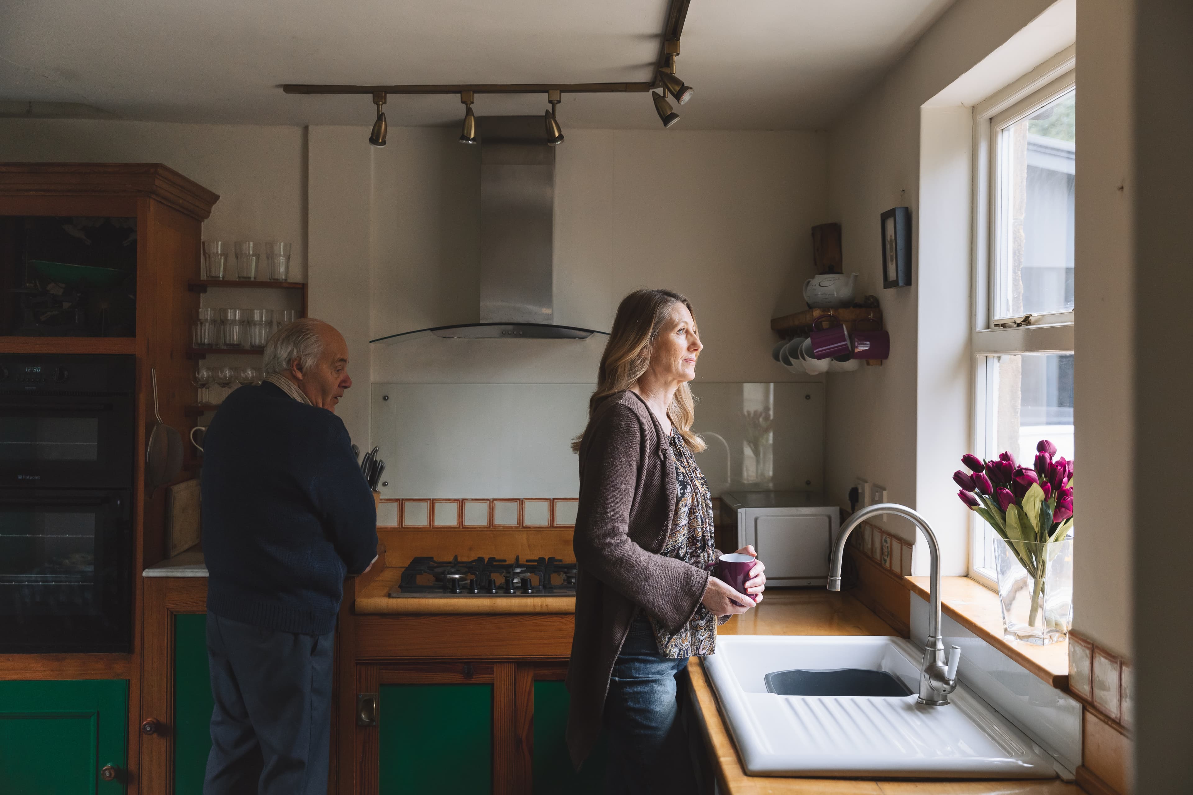 A woman looks out a kitchen window holding a mug, while an older man stands at the counter behind her. - Home Instead