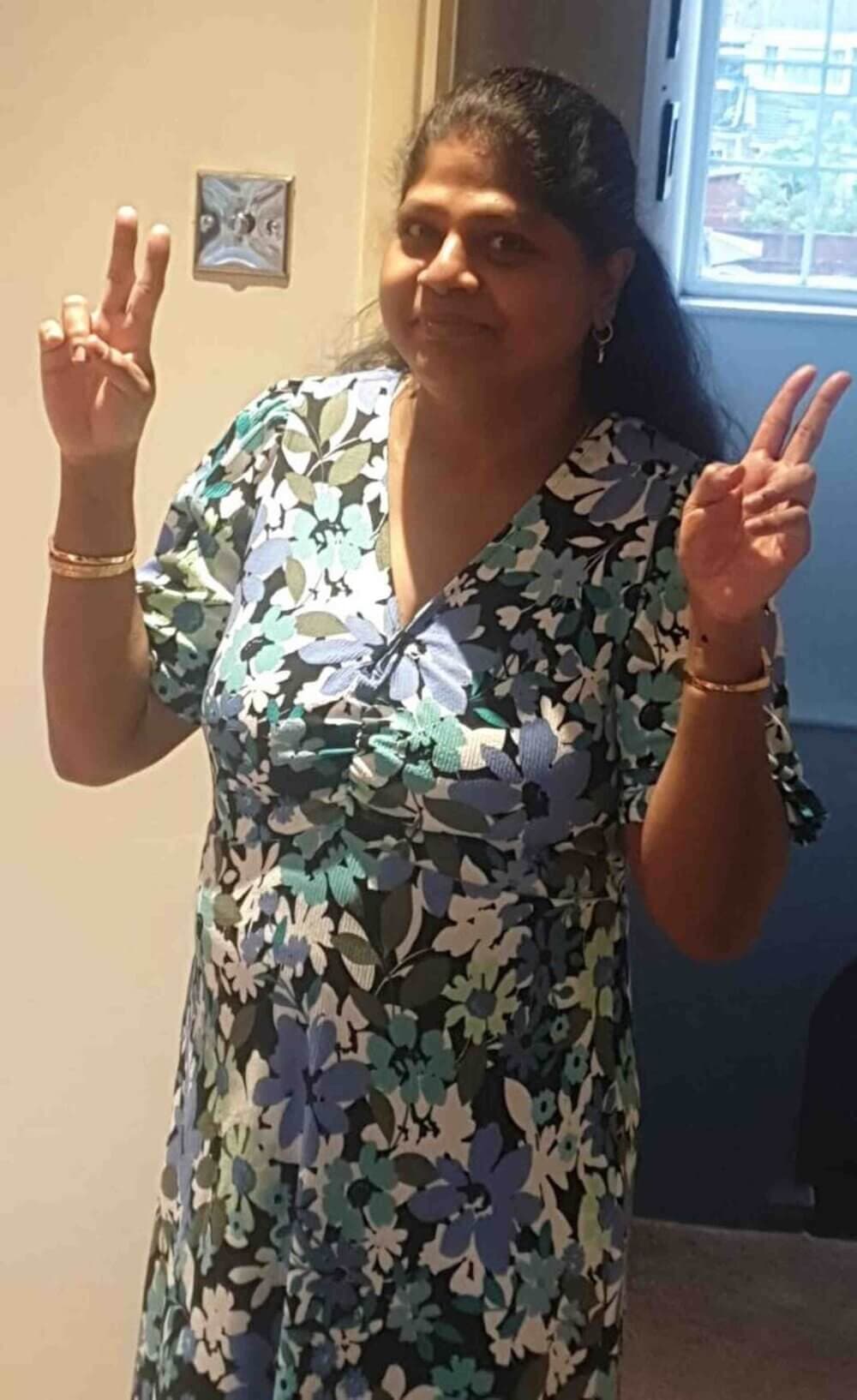 A person in a floral dress stands indoors, smiling and holding up two peace signs with both hands. - Home Instead