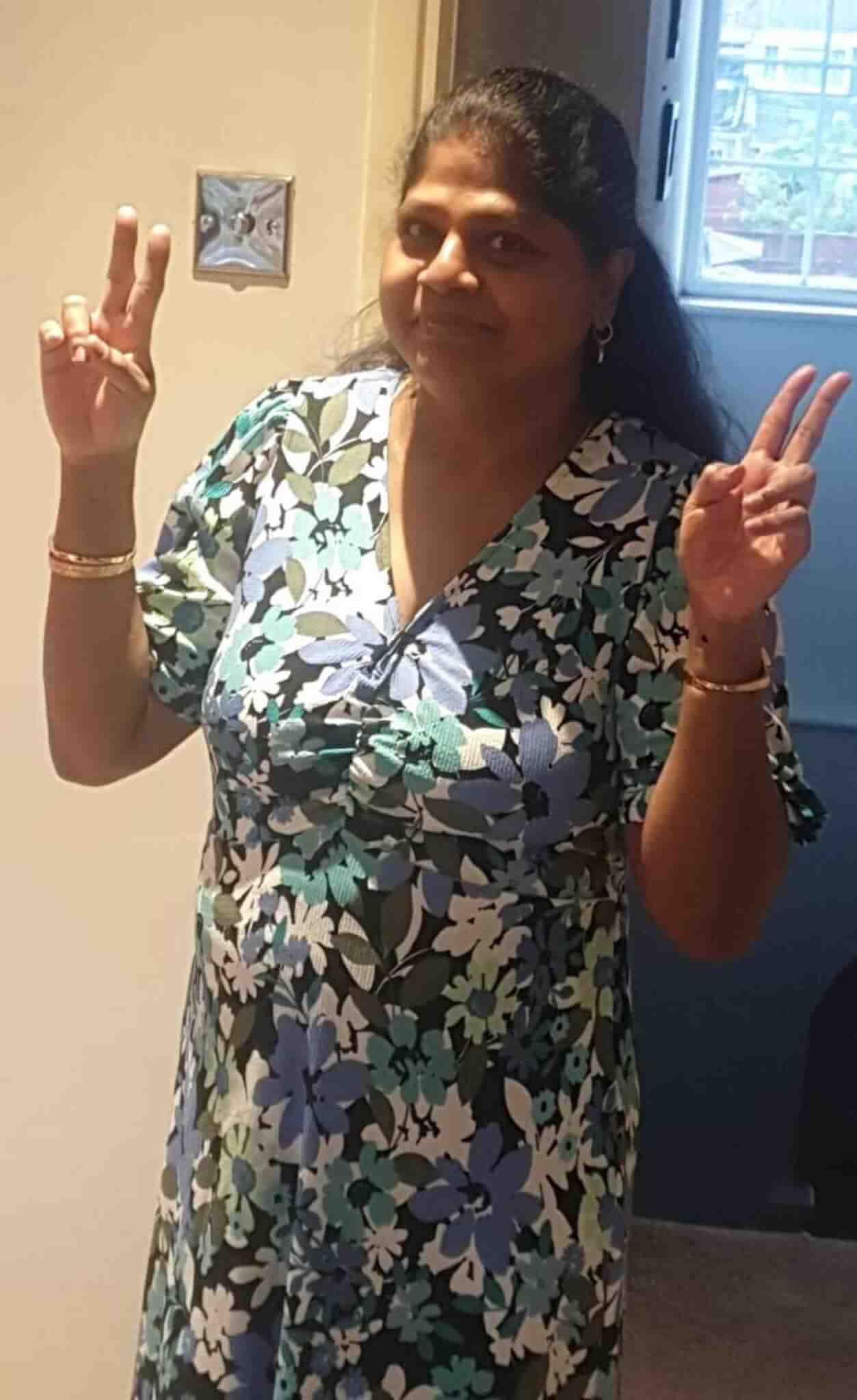 A person in a floral dress stands indoors, smiling and holding up two peace signs with both hands. - Home Instead