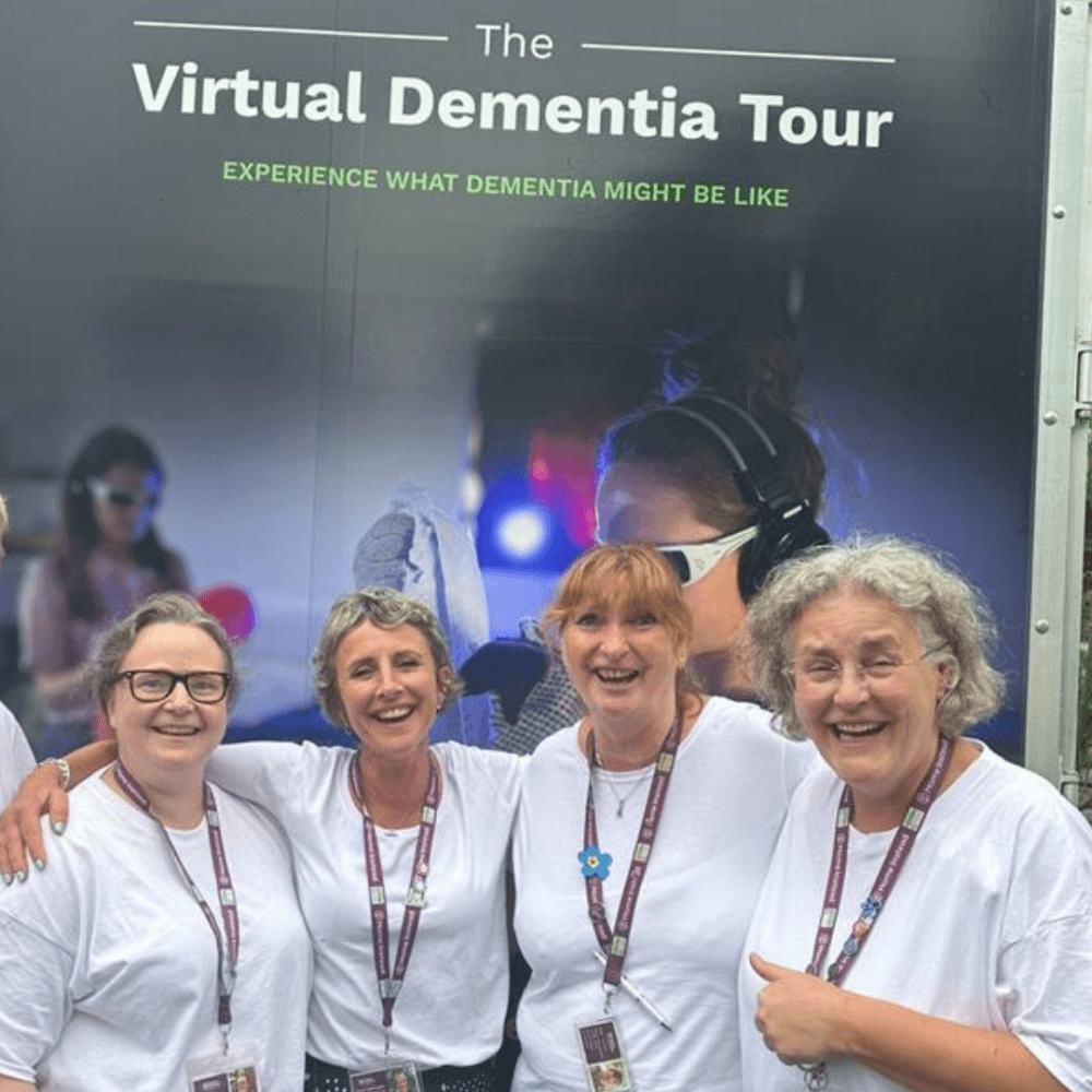 Four smiling women in white shirts stand together in front of a sign for "The Virtual Dementia Tour. - Home Instead