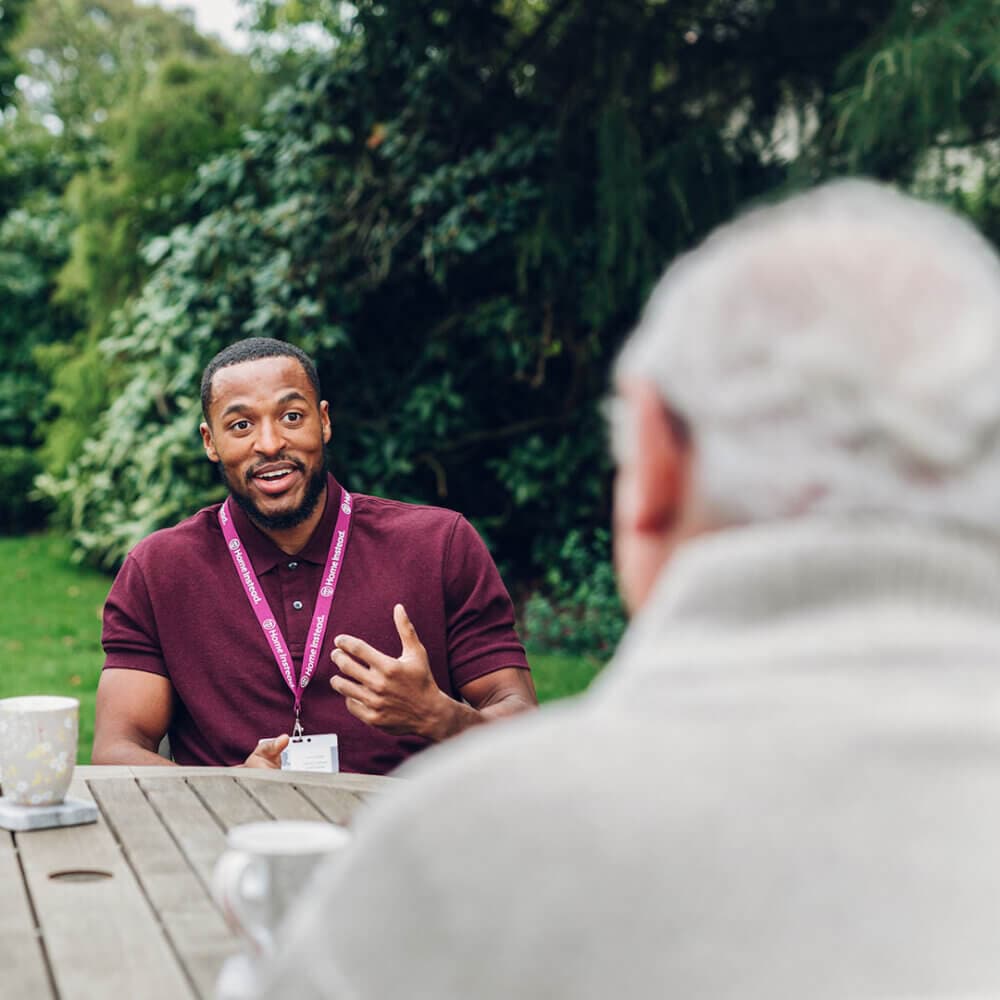 A man in a burgundy shirt talks to an elderly person across a table outdoors. They are seated in a garden area. - Home Instead