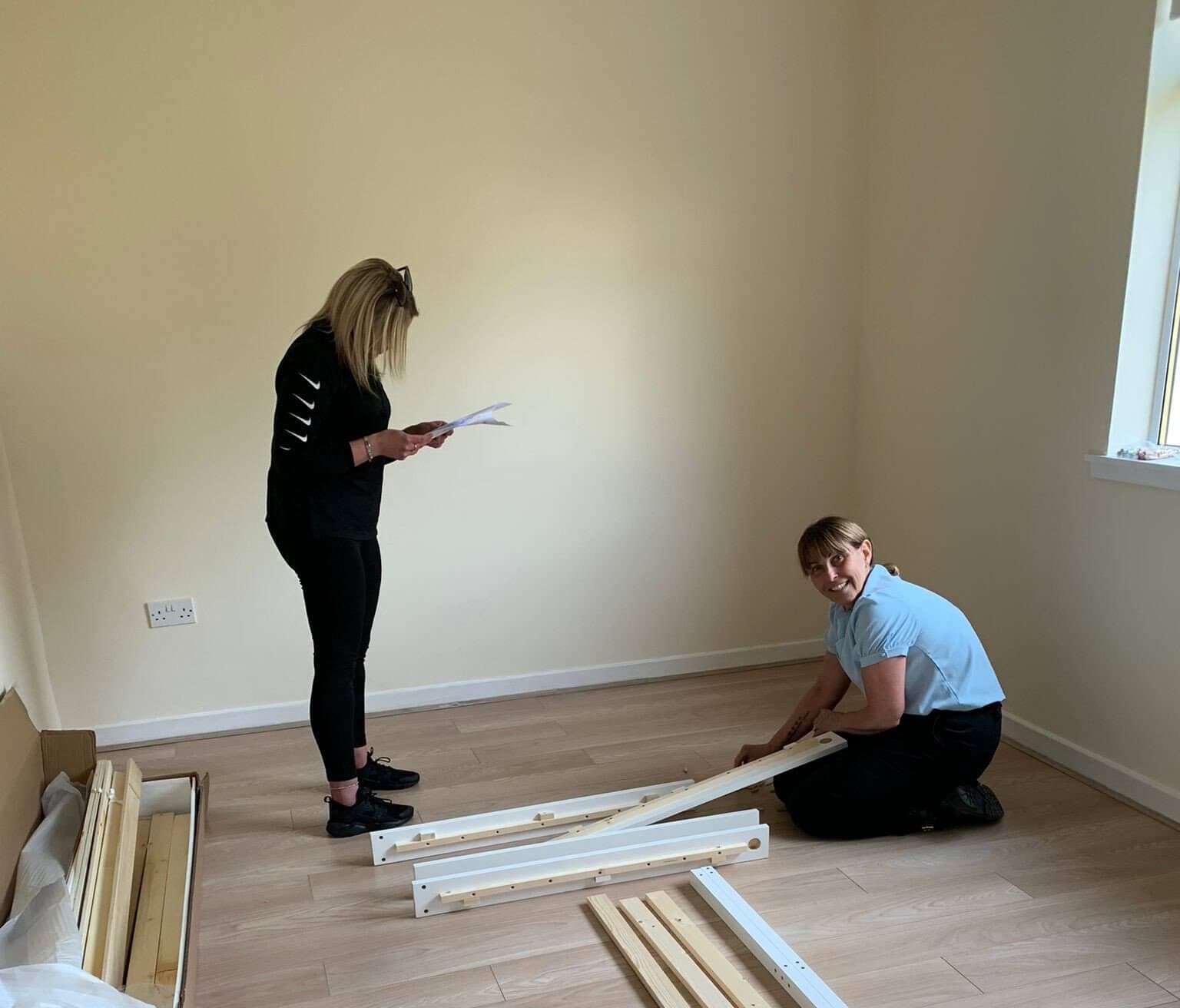 Two women assembling furniture in a room with light-colored walls and floor. One reads instructions; the other is on the floor. - Home Instead