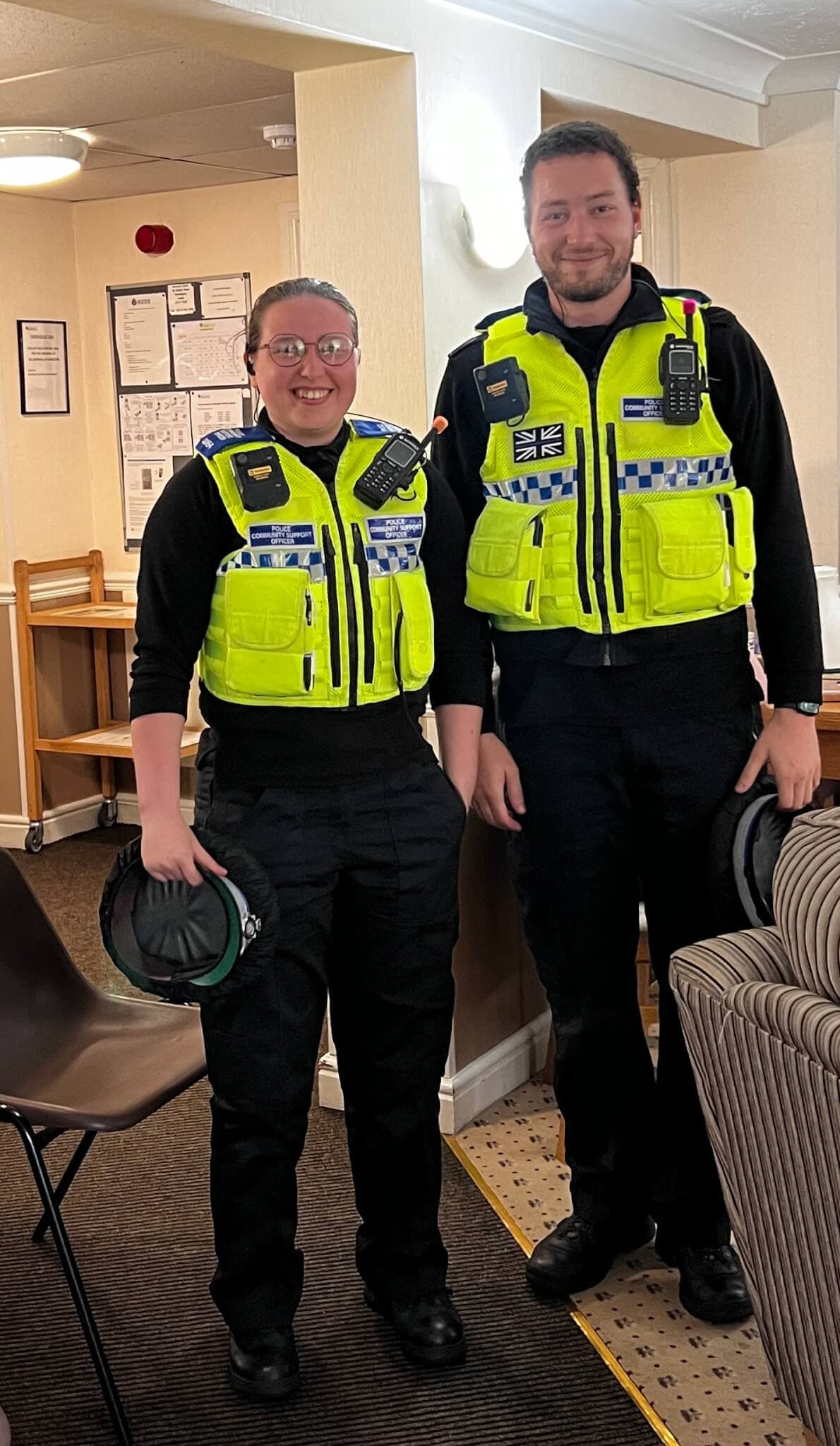Two police officers in high-visibility vests are standing indoors, smiling at the camera. - Home Instead