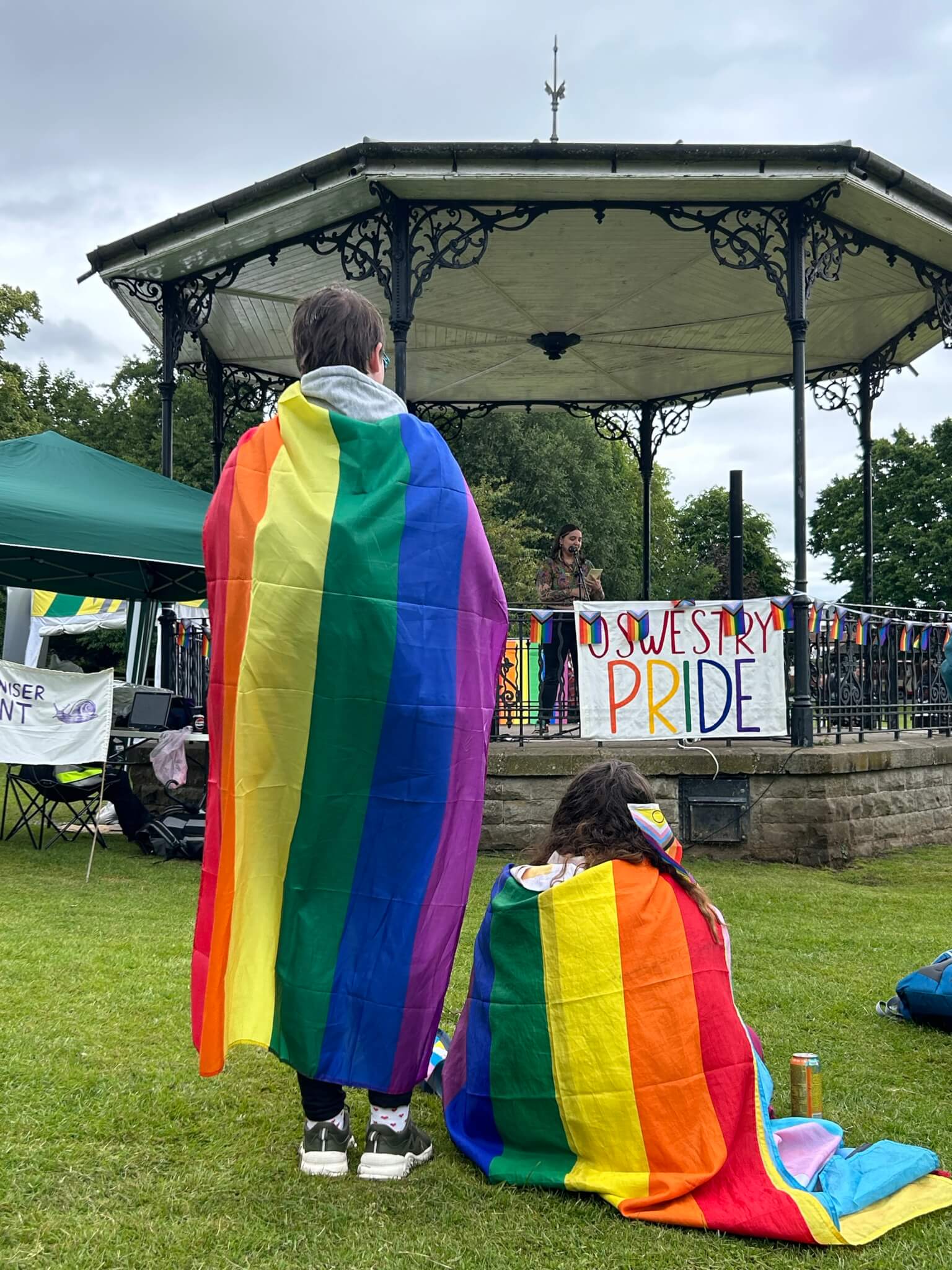 Two people draped in rainbow flags stand and sit in front of a gazebo at Oswestry Pride event. - Home Instead