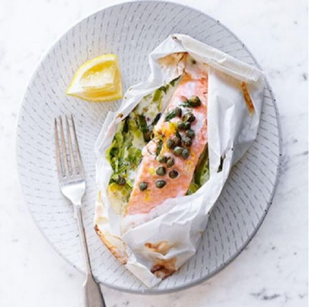 Baked salmon with capers and vegetables wrapped in parchment paper on a plate with a lemon wedge and fork. - Home Instead