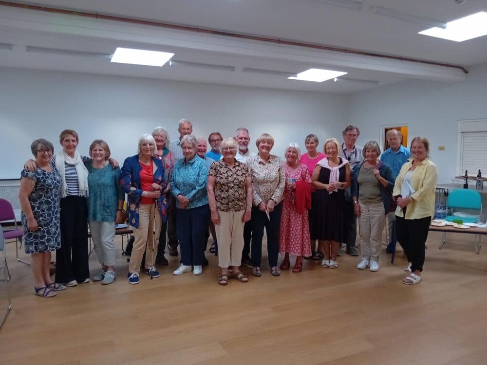 A group of people stands together inside a room, posing for a photo. They appear to be smiling and cheerful. - Home Instead