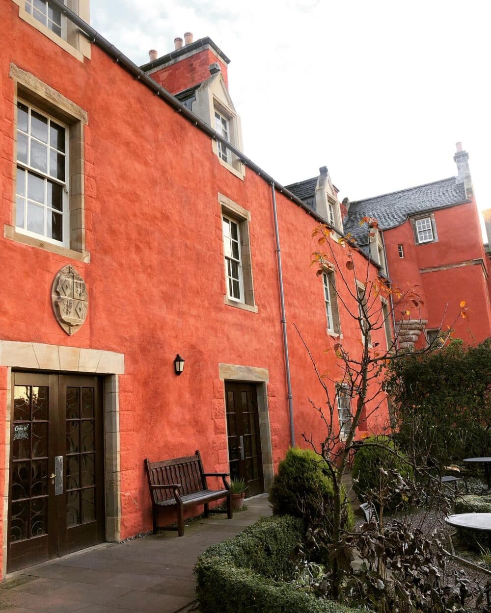 Historic building with red walls, small windows, a wooden bench, and a courtyard with several greenery and a leafless tree. - Home Instead