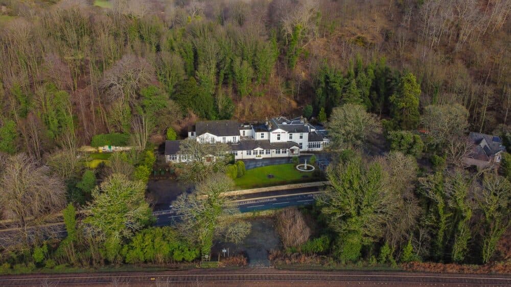 Aerial view of a large house surrounded by dense forest with a road and railway tracks visible at the bottom. - Home Instead