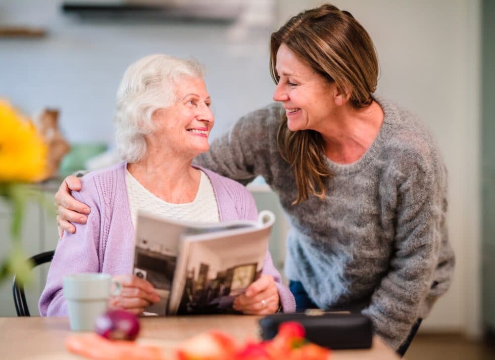 An elderly woman holding a magazine smiles at a woman standing beside her, both sharing a joyful moment in a cozy kitchen. - Home Instead