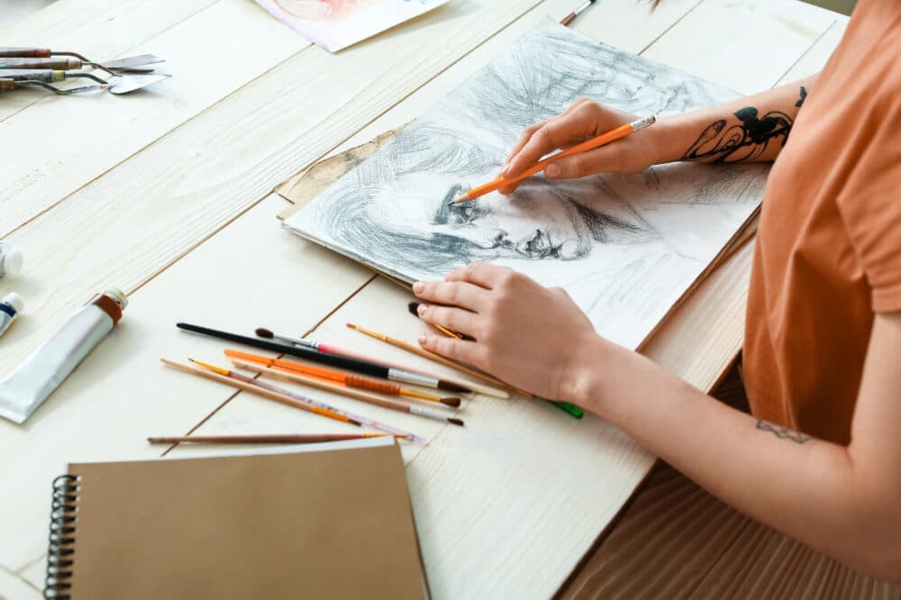 Person drawing a portrait with pencils, surrounded by art supplies on a wooden table. - Home Instead
