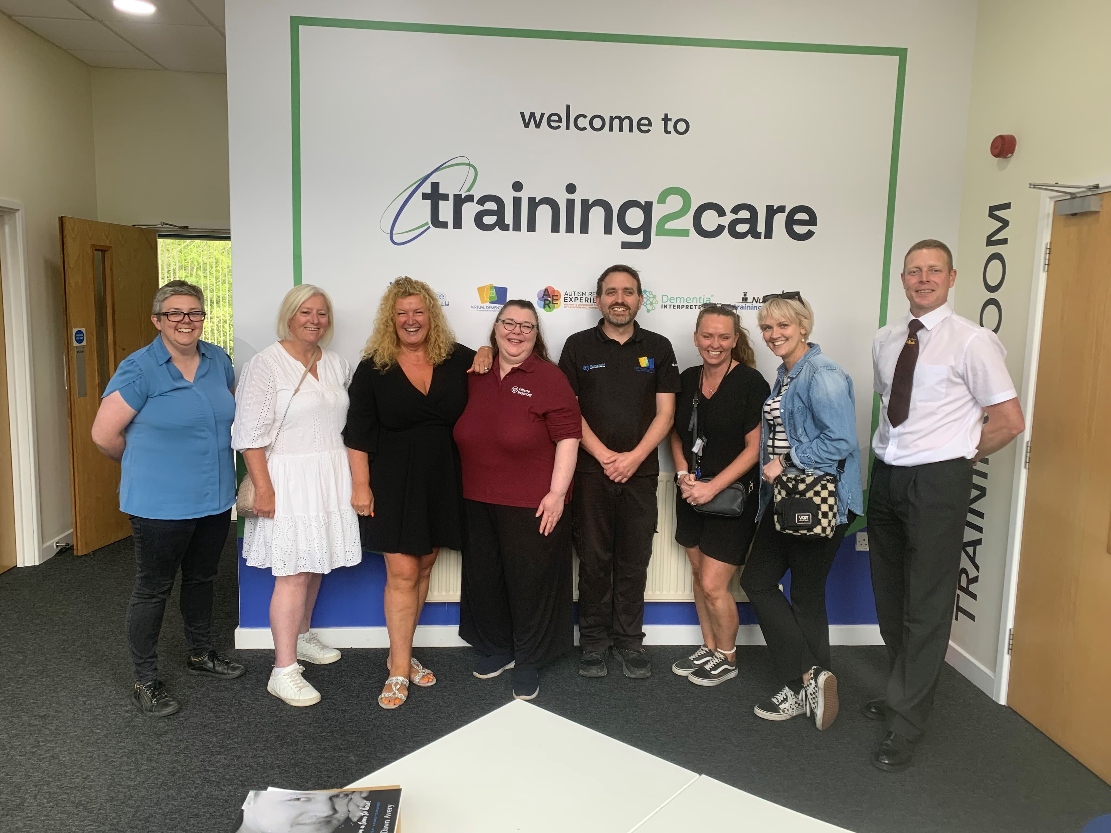 A group of eight people standing and smiling in front of a "Welcome to training2care" sign. - Home Instead