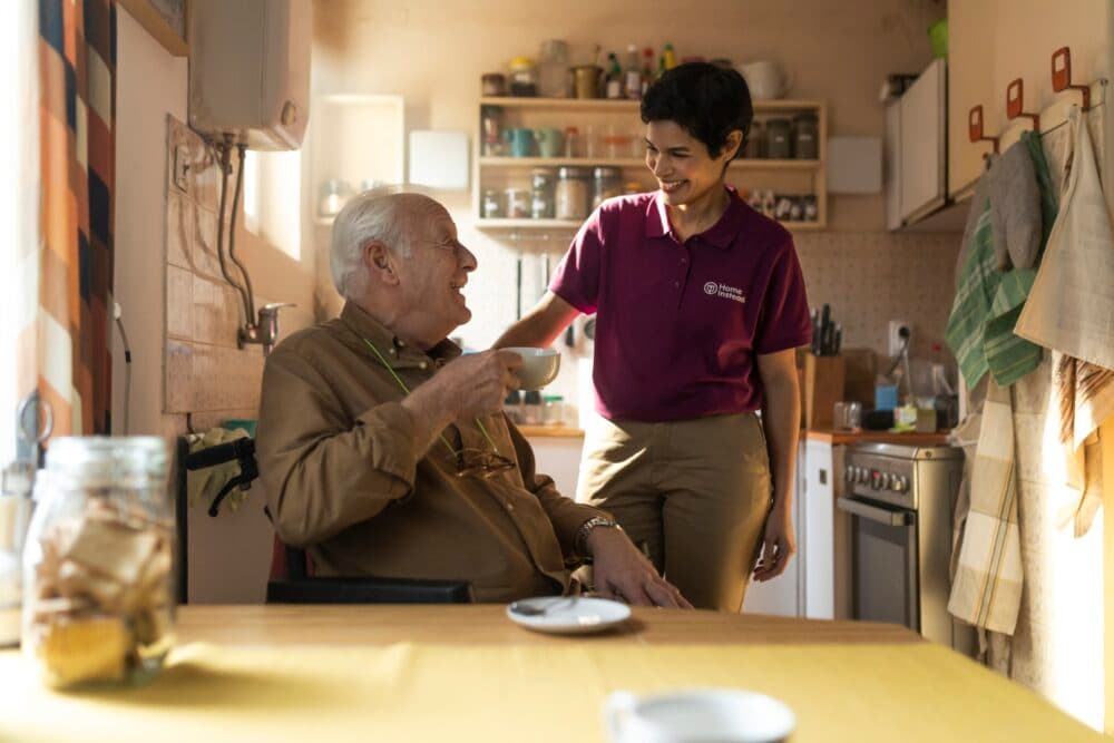 A smiling caregiver in a purple shirt helps an elderly man in a kitchen as he holds a cup of coffee. - Home Instead