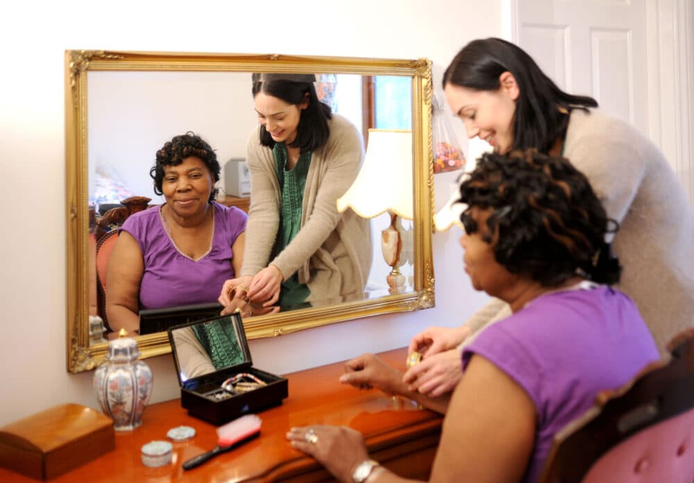 Two women, one sitting and one standing, look at jewelry on a vanity table in front of a large mirror. - Home Instead