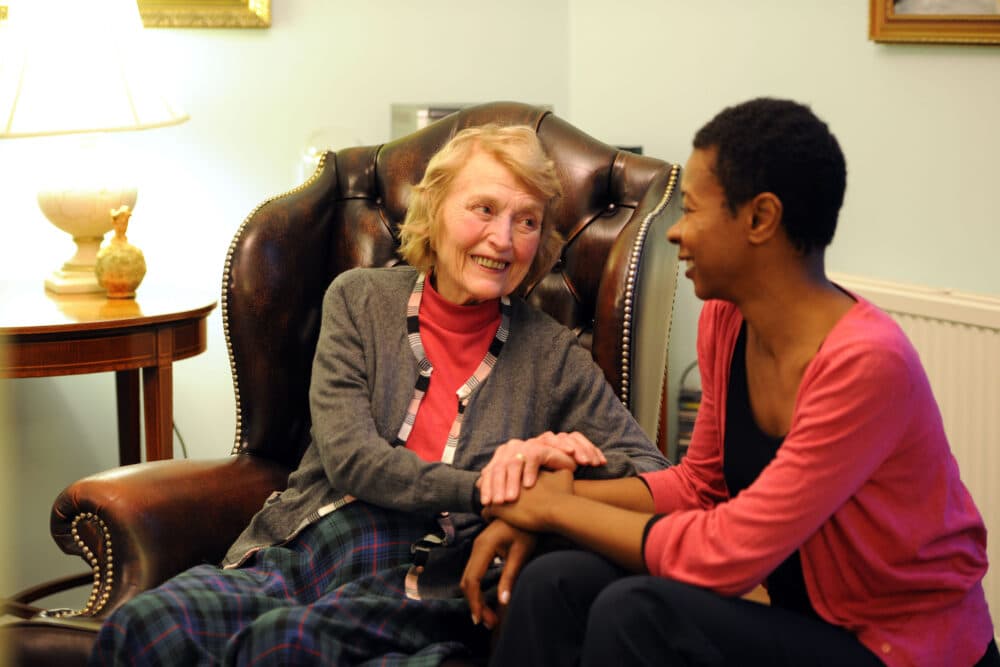 Two women share a warm moment, smiling and holding hands, while seated in a cozy living room with soft lighting. - Home Instead