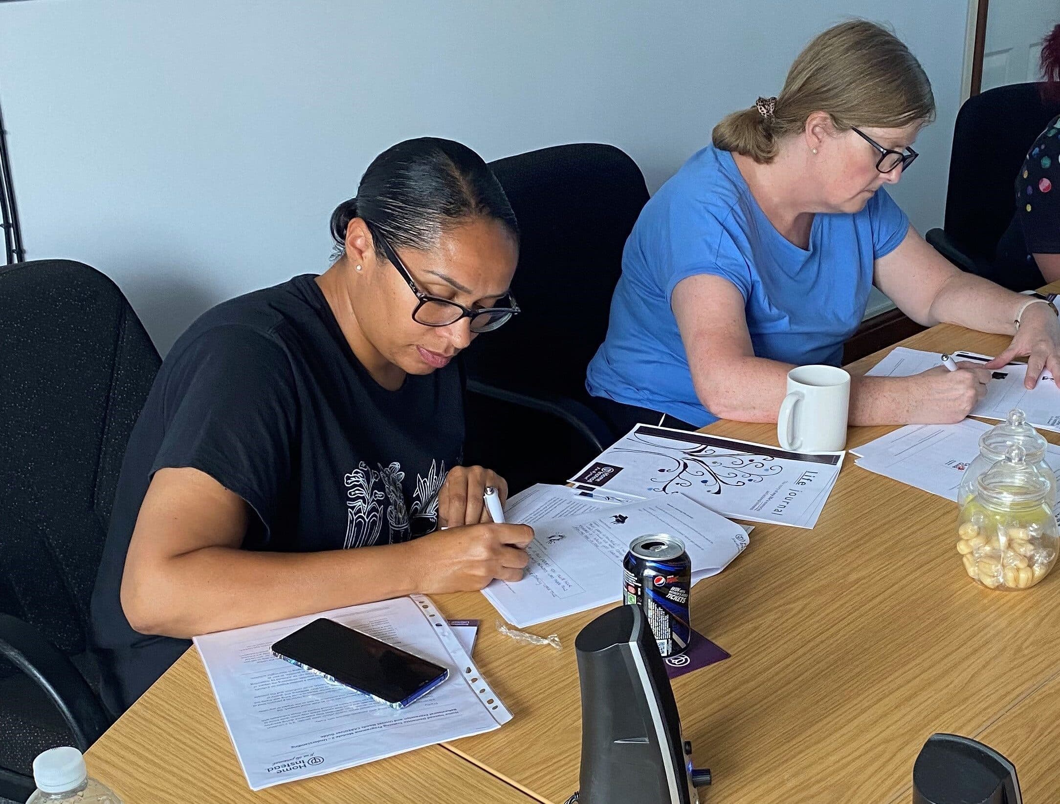 Two women sit at a table, focused on writing. Papers, a phone, and a drink can are on the table around them. - Home Instead