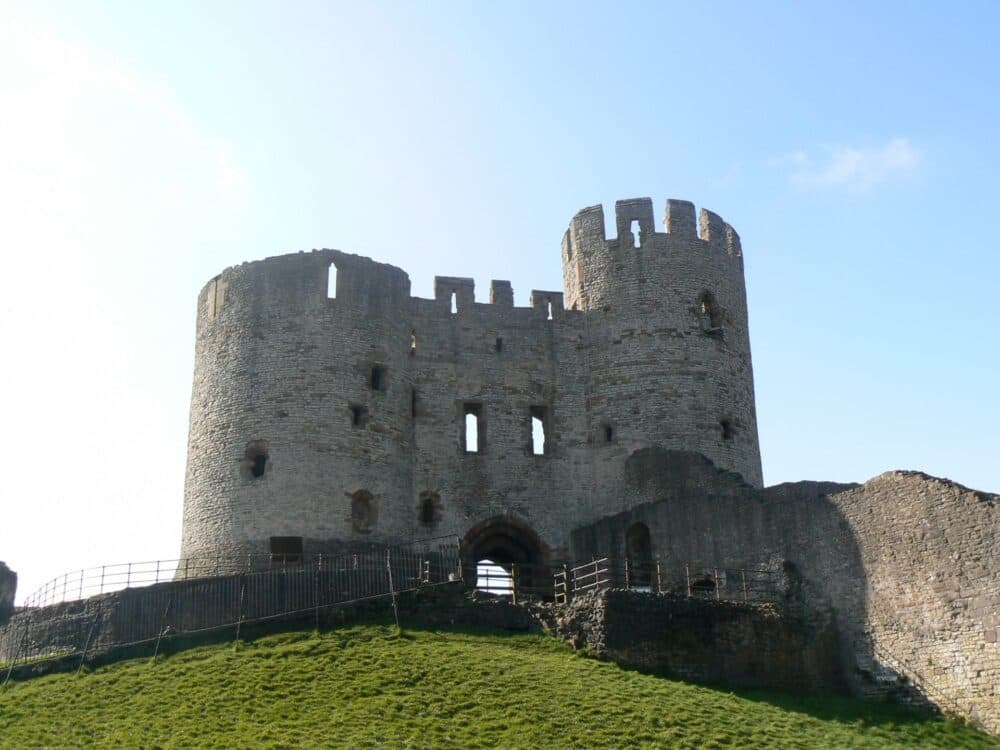 Ancient stone castle with round towers and arched entrance, situated on a grassy hill under a clear blue sky. - Home Instead