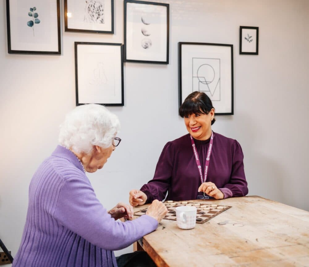 An elderly woman and a young woman play a board game at a wooden table with framed art on the wall behind them. - Home Instead