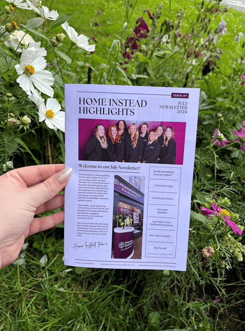 A hand holding a "Home Instead Highlights" newsletter with flowers and grass in the background. - Home Instead