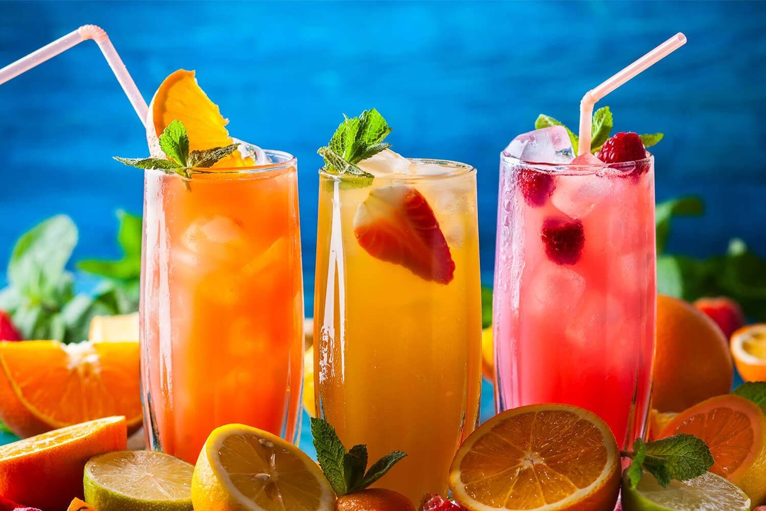 Three tall glasses of colorful fruit drinks with ice, garnished with mint, surrounded by assorted citrus fruits. - Home Instead