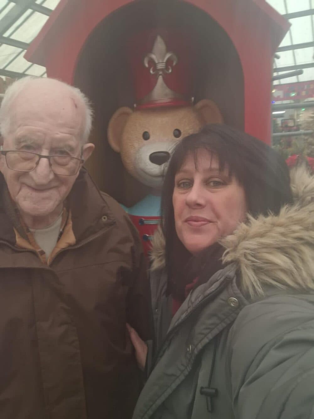 An elderly man and a woman take a selfie in front of a teddy bear statue dressed as a soldier. - Home Instead
