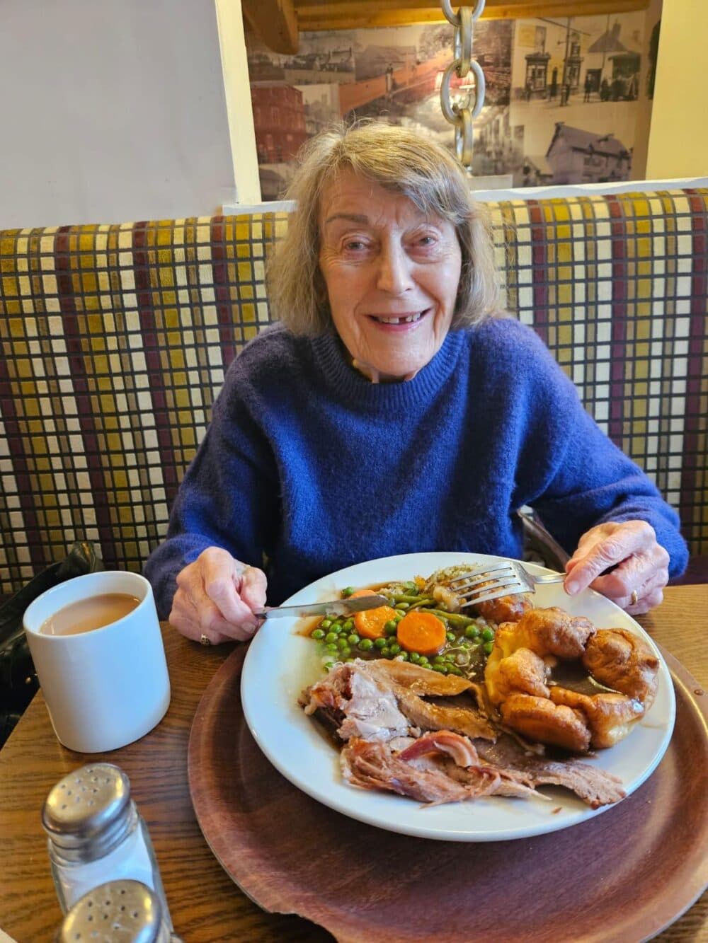 An elderly woman smiles, sitting at a table with a large plate of food and a cup of tea. - Home Instead