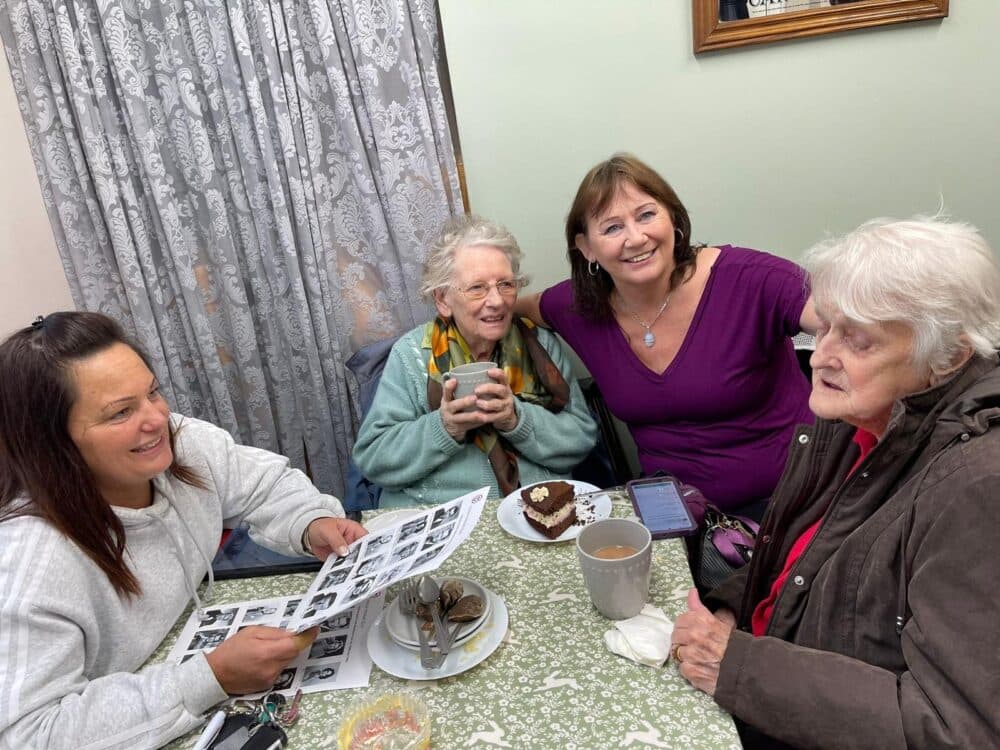 Four women gathered around a table, smiling and chatting with tea, cake, and photos in a cozy indoor setting. - Home Instead