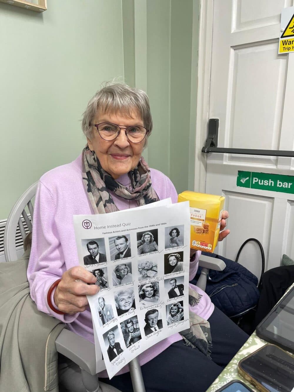 An elderly woman holding a quiz sheet and a box of tea, smiling. She is seated in a room with green walls. - Home Instead