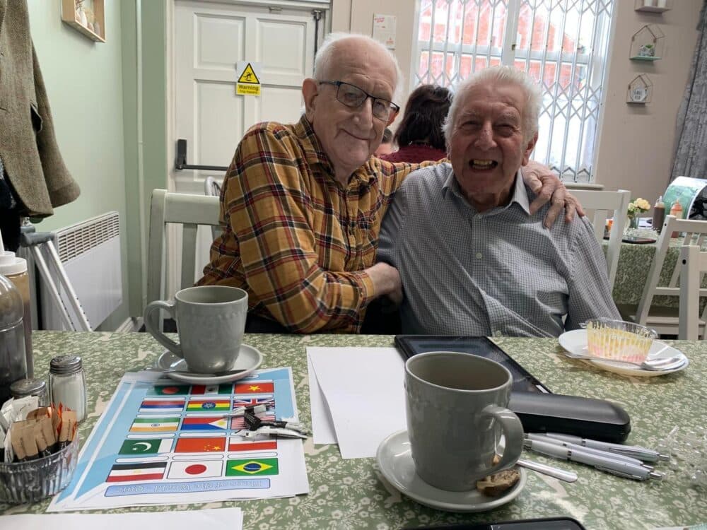Two elderly men smiling and sitting at a table with coffee mugs and a flag worksheet in a cozy room. - Home Instead