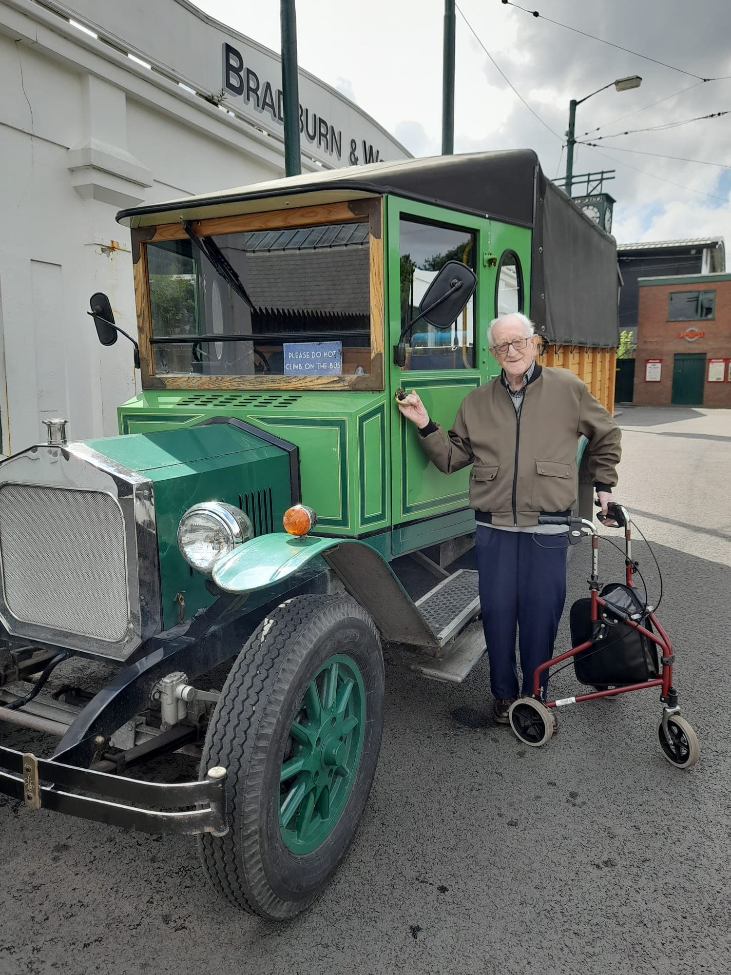 An elderly man, standing with a walker, poses beside a green vintage truck on a sunny day near a building. - Home Instead