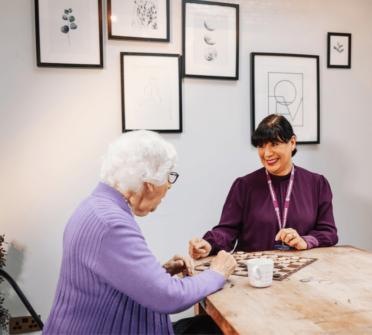An elderly woman and a caregiver play a game at a table in a decorated room with framed art on the walls. - Home Instead