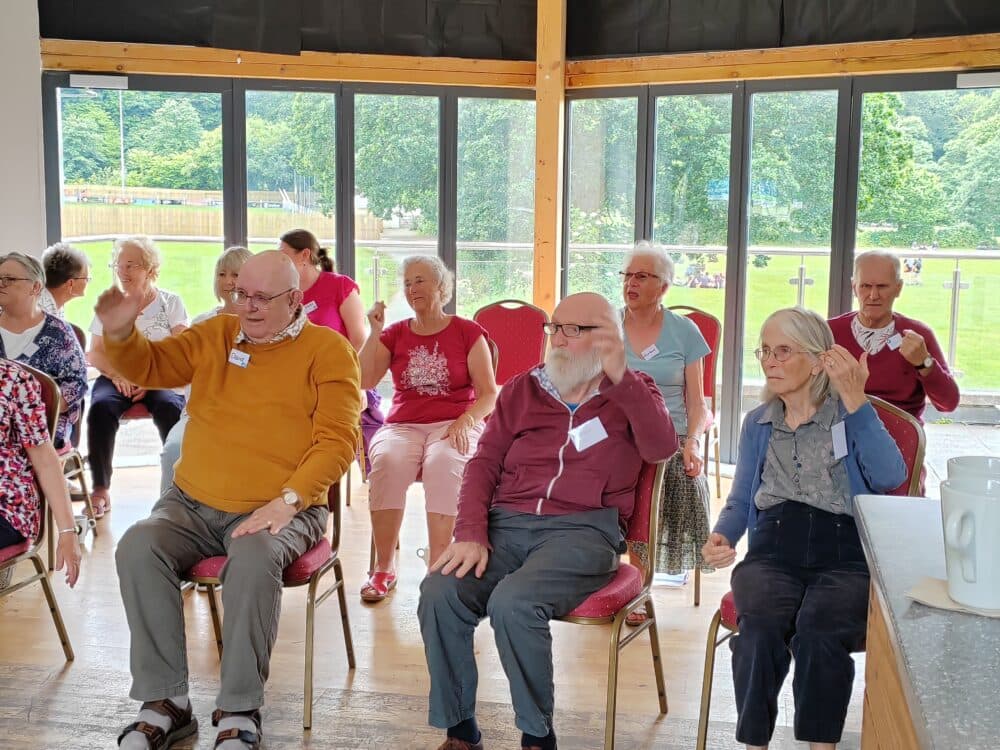 A group of elderly people sitting on chairs, engaging in activities inside a room with large windows. - Home Instead