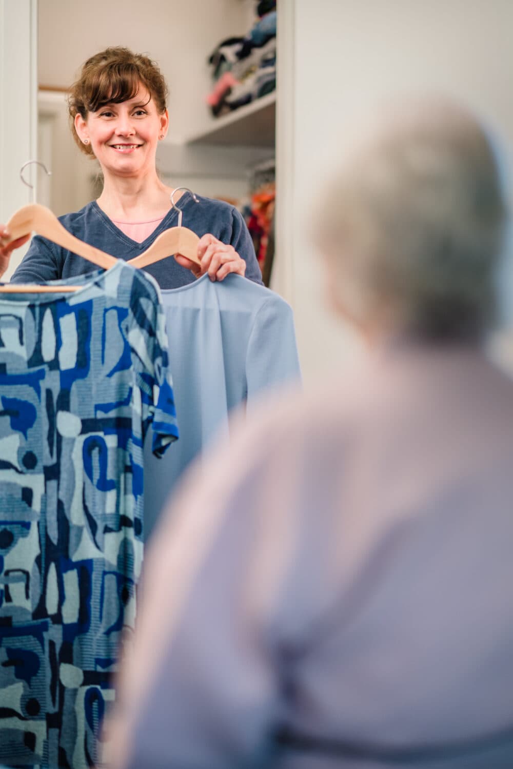 A smiling woman holds two outfits on hangers, showing them to another person whose back is turned. - Home Instead