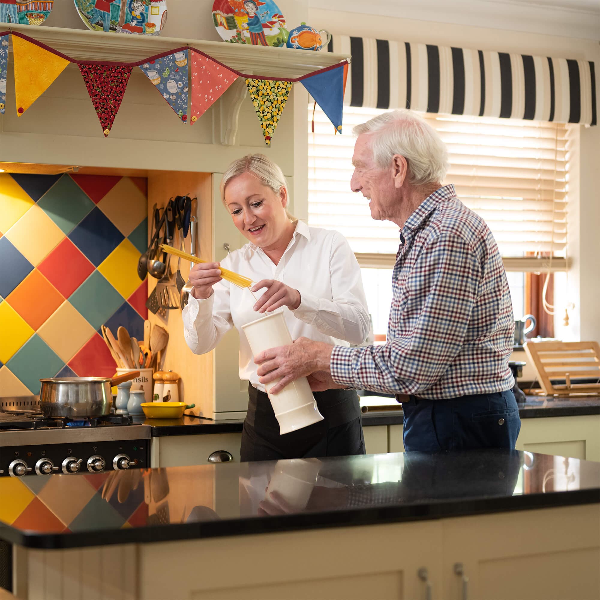 A woman helps an elderly man pour pasta into a pot in a colorful kitchen. Bright bunting hangs above. - Home Instead
