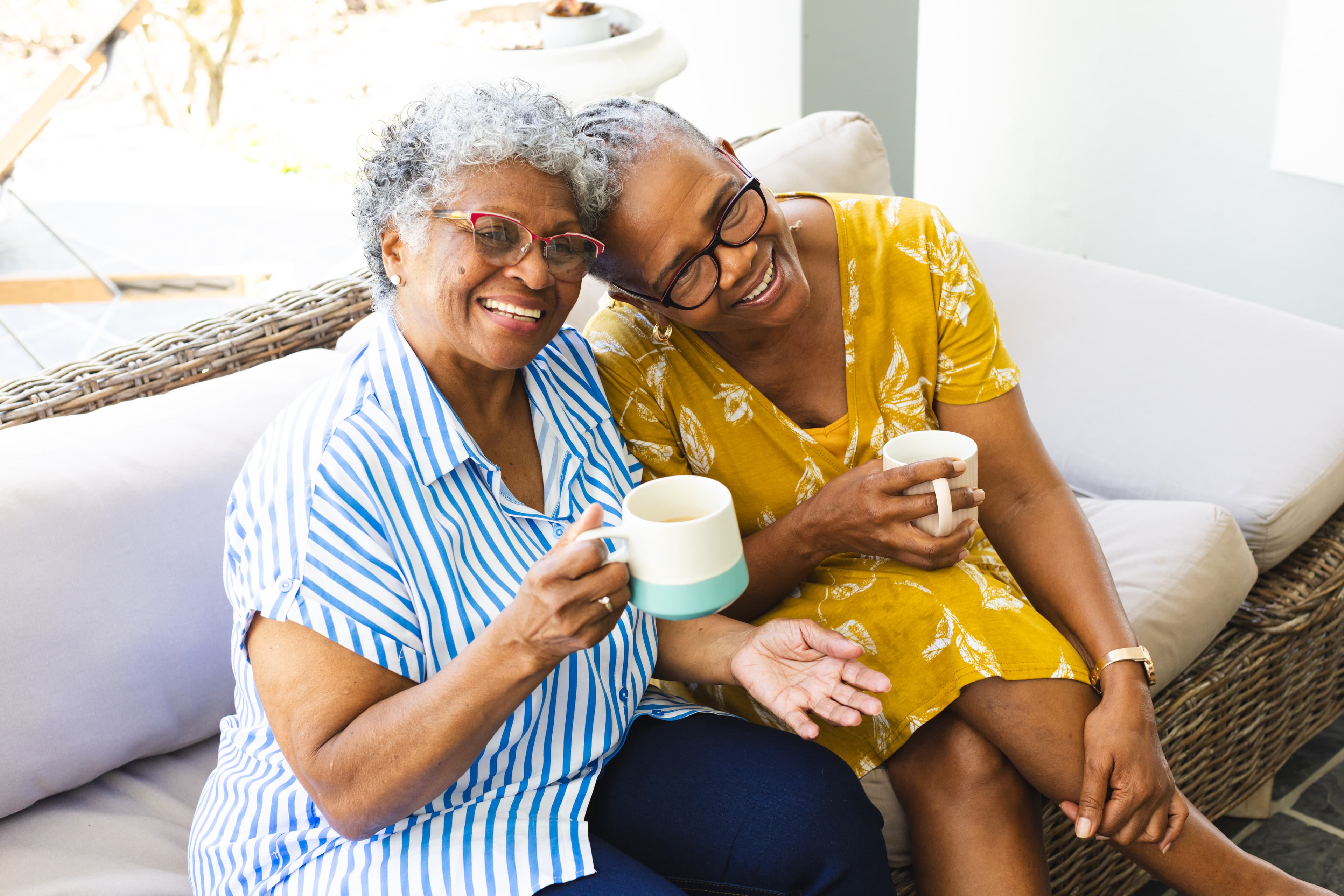 Two elderly women smiling while sitting on a couch, holding mugs, and enjoying a cozy moment together. - Home Instead