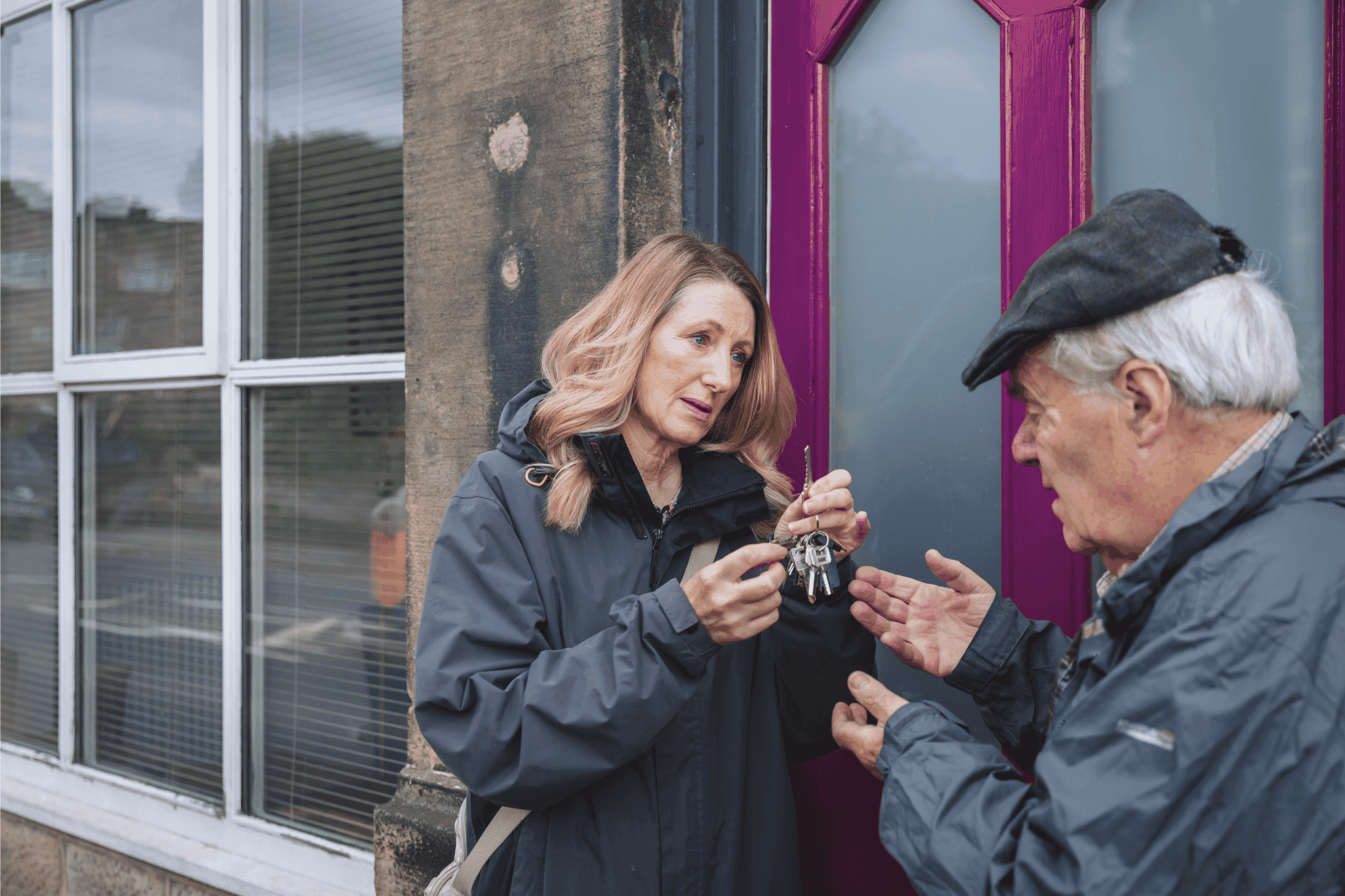A woman hands a set of keys to an older man in front of a building with large windows and a purple door. - Home Instead