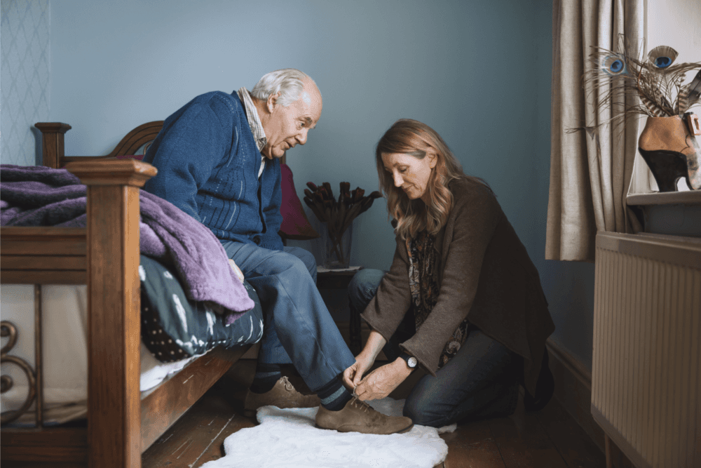 A woman kneels to help an elderly man tie his shoes by a bed in a warmly lit room. - Home Instead
