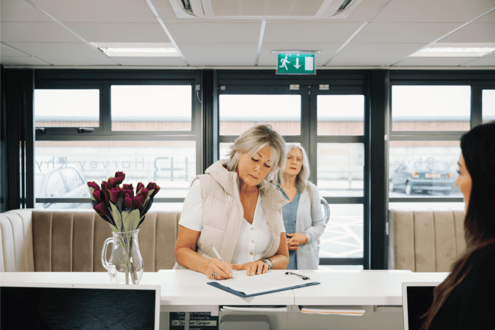 A woman signs a document at a reception desk, with another woman standing behind her. A vase of flowers is on the desk. - Home Instead