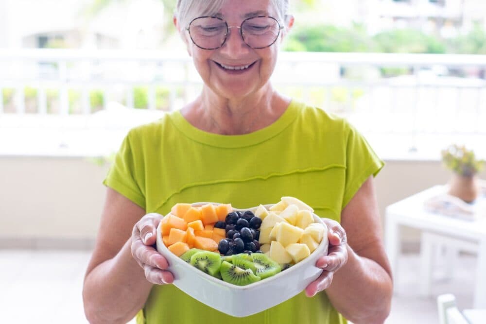 Smiling woman holding a bowl of mixed fruits including cantaloupe, kiwi, blueberries, and apple slices. - Home Instead