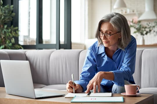 Elderly woman with glasses writing in notebook, seated on couch near laptop and cup of coffee. - Home Instead