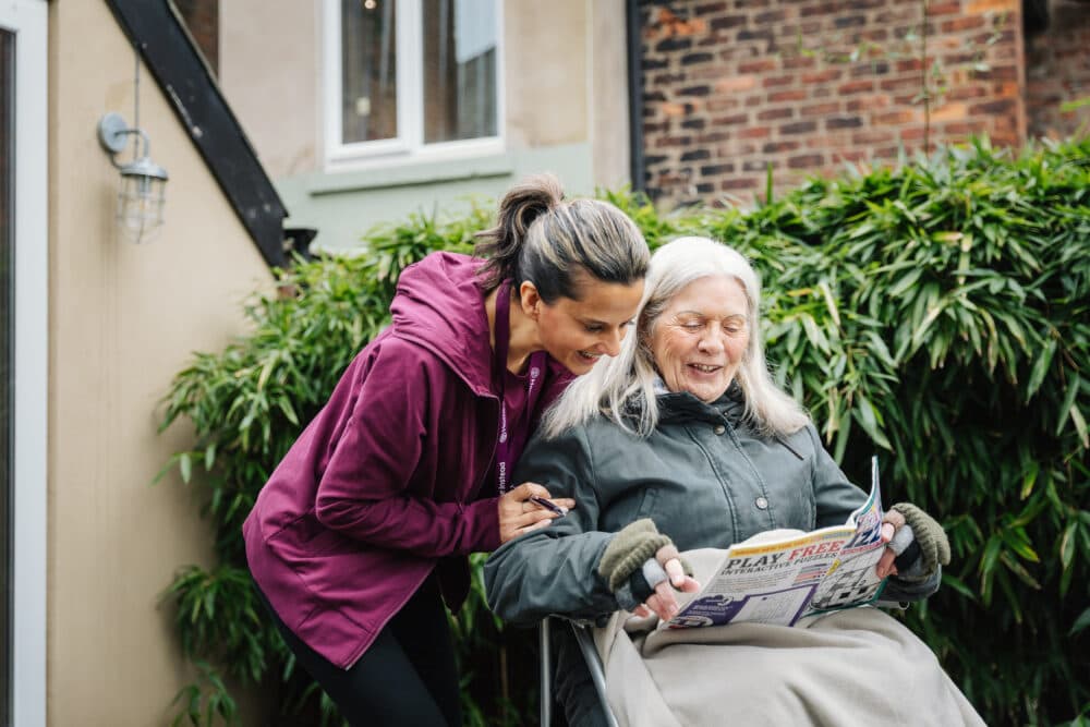 Caregiver smiling and leaning over elderly woman reading a newspaper in a wheelchair outdoors. - Home Instead